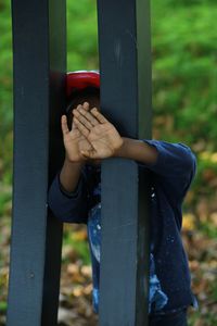 Boy hiding face through metallic fence on field