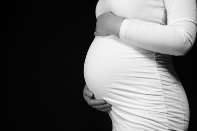 Midsection of pregnant woman touching abdomen and standing against black background 