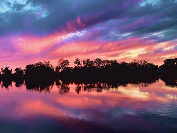 Silhouette trees by lake against dramatic sky during sunset