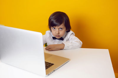 Serious boy child businessman in a white shirt and bow tie sits at a laptop desk in a yellow office