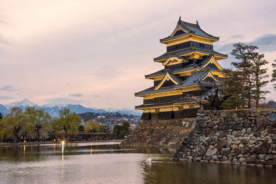 Traditional building by lake against sky during sunset