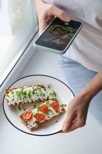 Woman takes a photo on cellphone hold plate with rye crisp bread with creamy cheese and micro greens