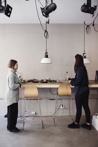 Full length of mid adult female colleagues standing by food on table against wall at workshop