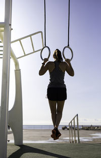 Full length rear view of woman hanging from gymnastic rings against sky