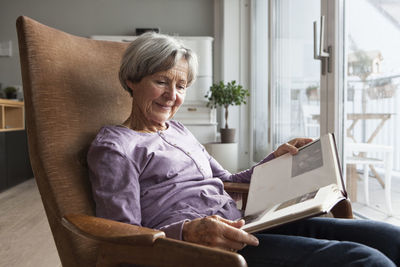 Portrait of senior woman sitting on armchair at home watching photo album