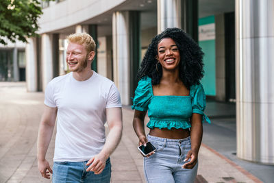 Smiling african american woman in blue top and jeans with afro hairstyle and blond haired man in white t shirt walking along paved sidewalk and chatting while looking at camera