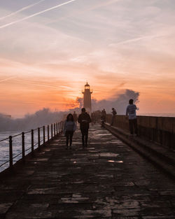 People by lighthouse amidst sea against sky during sunset