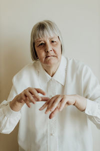 Sad elderly woman in white blouse gesturing with hands on light background looking at camera