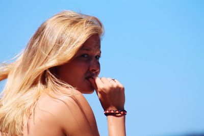 Young woman nail biting while looking away against clear blue sky