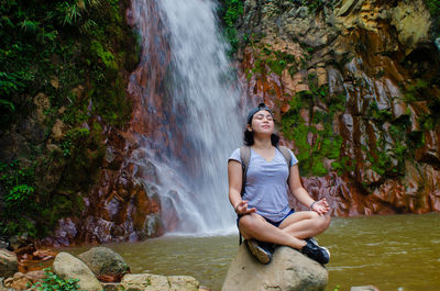 Young woman mediating while sitting on rock against waterfall in forest