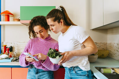 Young woman sharing mobile phone with female friend while leaning on kitchen counter at home