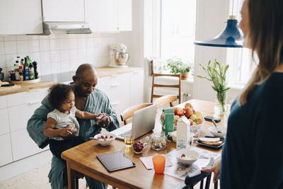 Woman looking at man using laptop while carrying daughter during breakfast at table