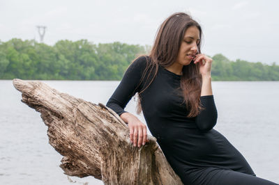 Beautiful young woman sitting on wood against trees