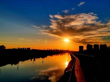 Silhouette buildings by river against sky during sunset