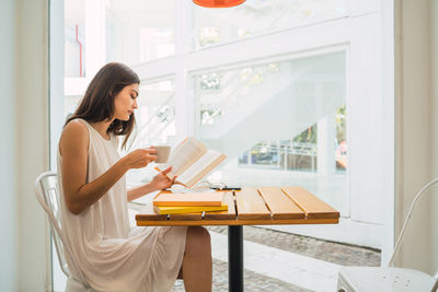 Side view of young woman enjoys coffee while reading book in cafe