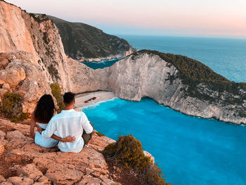 Rear view of woman sitting on rock by sea against sky