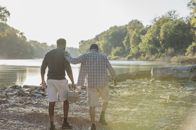 Rear view of male friends with fishing rods walking on rocks by lake