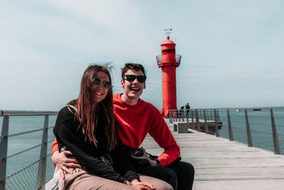 Friends sitting on railing by sea against sky