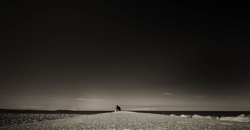 Distant view of man sitting by sea shore against sky