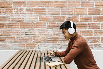 Side view of young man using mobile phone against brick wall