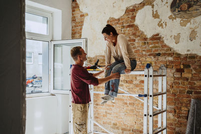 Man talking with woman sitting on ladder in front of brick wall during home renovation