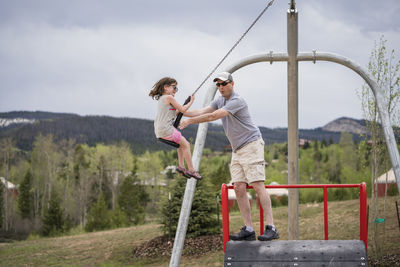 Father pushing daughter sitting on chain swing against cloudy sky at park