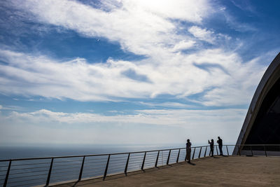 People standing on railing by sea against sky