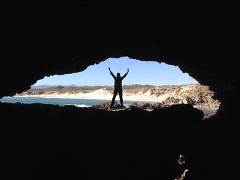 Silhouette man with arms raised at cave by sea