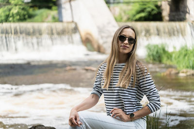 A woman in sunglasses, a striped top and white jeans near the river against the backdrop of the dam