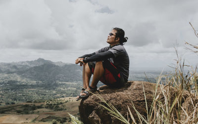 Young man sitting on mountain against sky