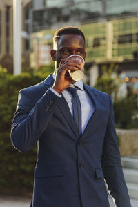Serious adult formally dressed african american male manager drinking takeaway coffee while standing against blurred urban background of modern city