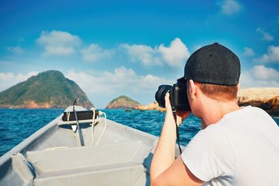 Side view of man photographing while traveling in boat on sea against blue sky
