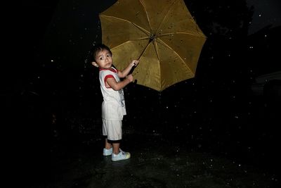Boy with umbrella looking away in rain while standing on road during night