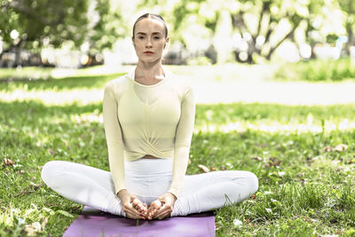 A young woman with long blonde hair does yoga in the summer in nature by the pond in the park.