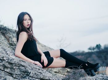 Full length portrait of young woman relaxing on rock against sky