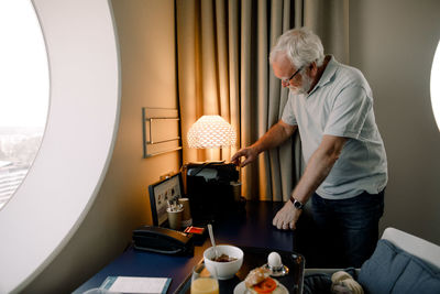 Senior man taking drink from espresso maker while standing by table in hotel room