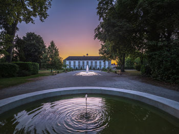 Fountain in swimming pool by lake against sky at sunset