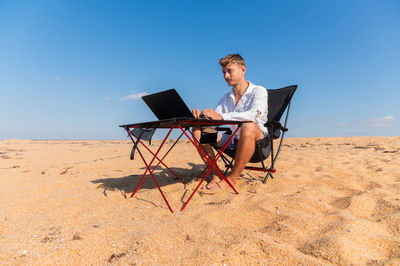 Side view of man using laptop at beach against clear blue sky