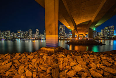 Illuminated bridge over river by buildings against sky at night