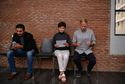 Business people reading documents while sitting on chairs against brick wall