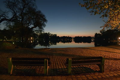 Empty bench by lake at park against sky at night