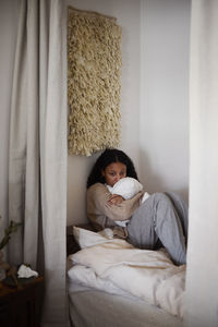 Pensive young woman sitting on bed and hugging pillow