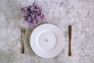 Plate with purple lilac flower, fork and knife on gray concrete background, top view.