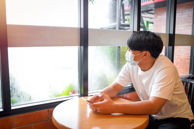 A handsome young teenage boy wear mask playing on smart mobile phone waiting in cafeteria. 