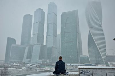 Rear view of man sitting against skyscrapers during snowfall