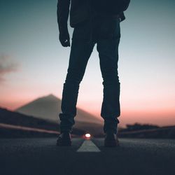 Low section of man standing on road against sky during sunset