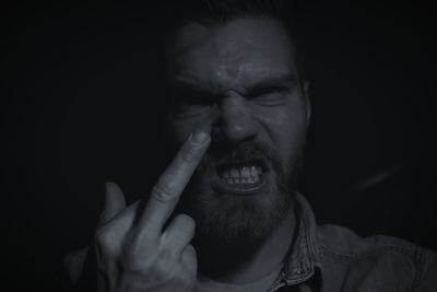 Close-up portrait of frustrated bearded man showing middle finger against black background