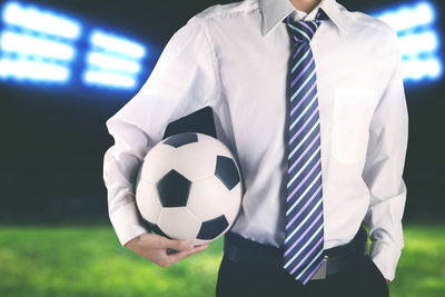 Midsection of businessman holding soccer ball on field at night