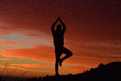 Silhouette man with arms raised standing against orange cloudy sky during sunset