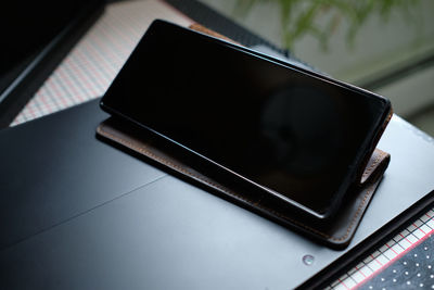 Black cellphone in a brown leather case is stood up on top of a black lap top with blank screen